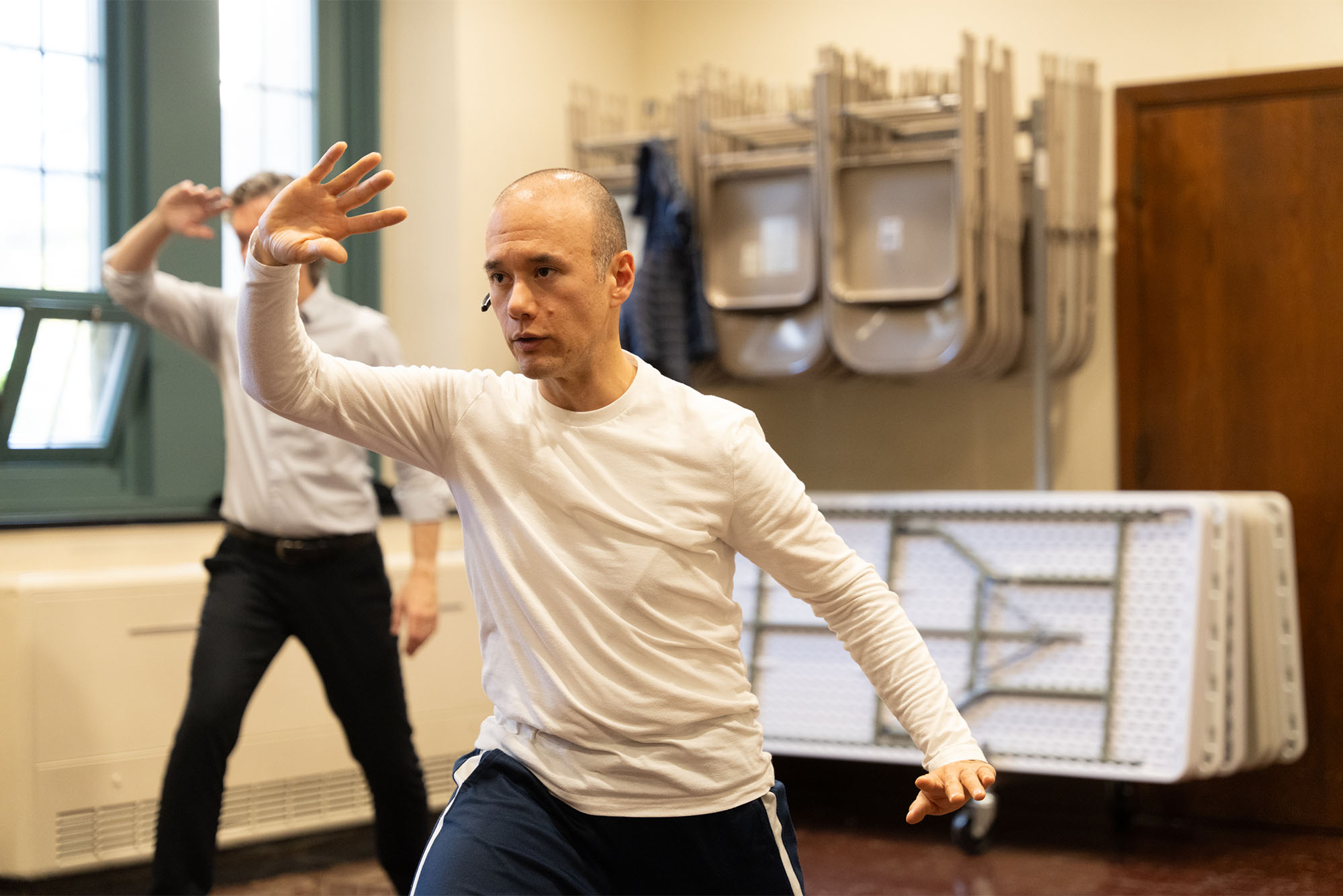 Photo: A man wearing headphones and casual workout attire guides a tai chi class with about 5-6 people following his movement