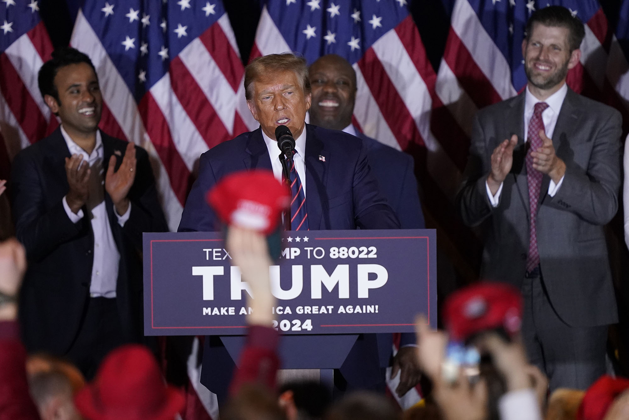 Photo: Republican presidential candidate former President Donald Trump speaks at a primary election night. He stands at a podium that reads TRUMP and an audience member reaches up to wave a red MAGA hat.