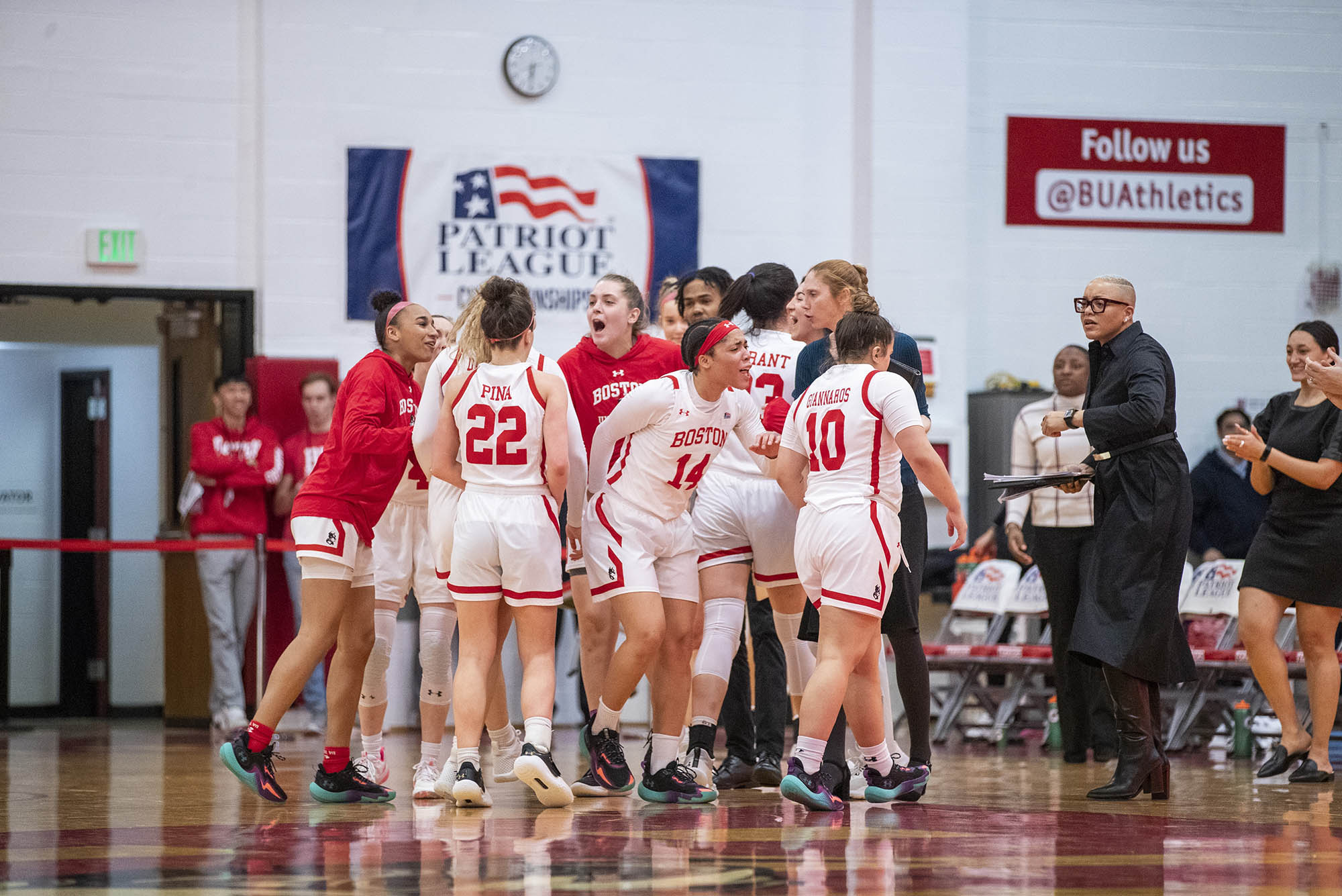 Photo: A photo of BU's Women's Basketball Team celebrating a great moment on the court. The team is huddled near the sidelines of the court. They are wearing their white uniforms with red detailing on the sides and for the numbers.