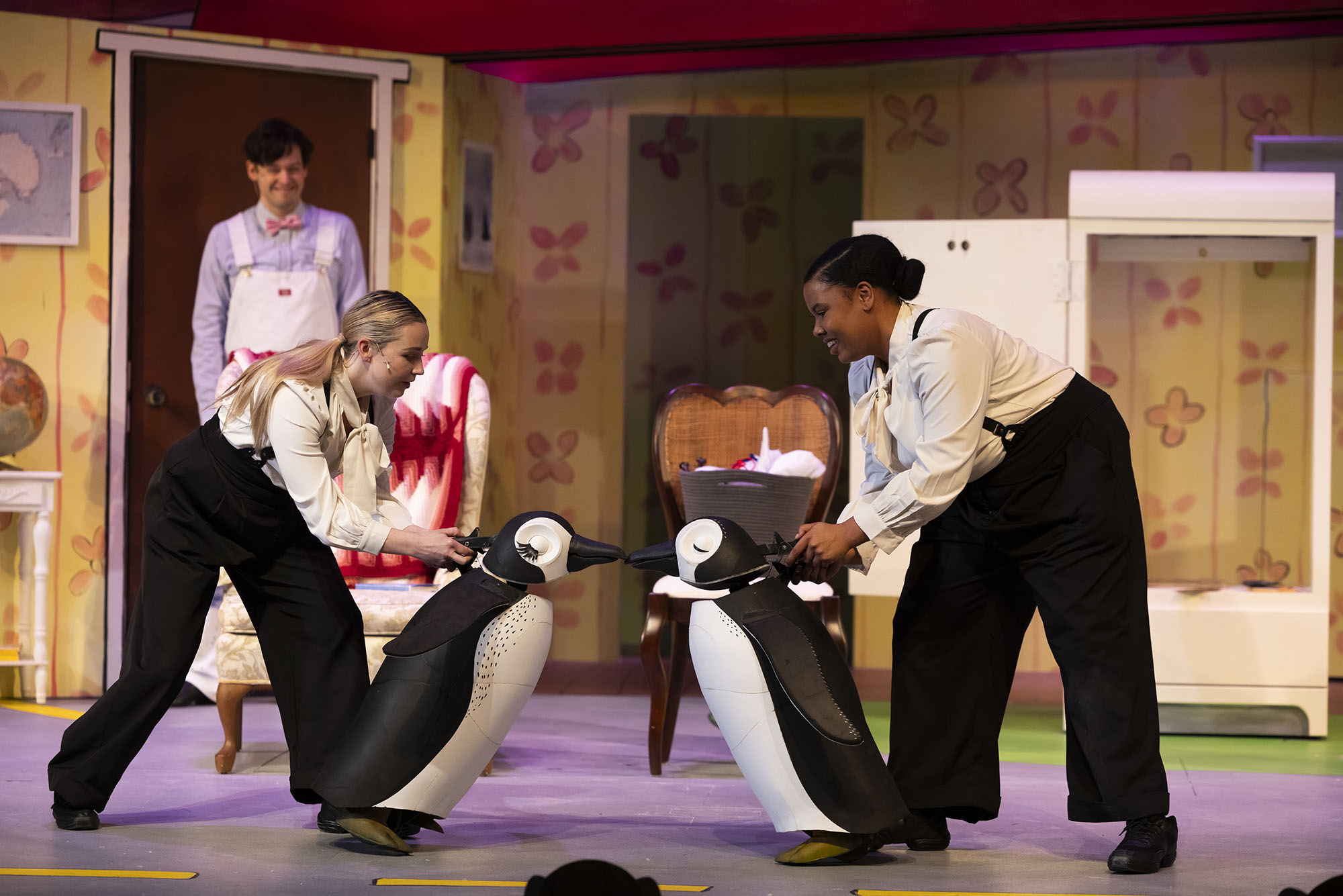 Photo: Two actors use a two prop penguins to mimic a "kiss" on stage. They background is colorful and comic like.