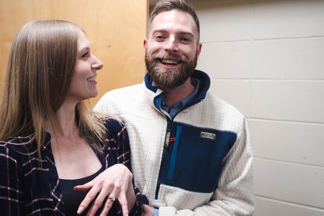 Photo: Two people, a white woman with blonde long hair and a white man with a blonde beard, celebrate as they just got engaged in the common room they met in.