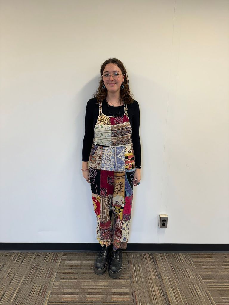 Photo: A photo of Victoria Witkowski, a young woman wearing platform docs, multi-patterned cotton overalls, a black long-sleeve, and glasses.
