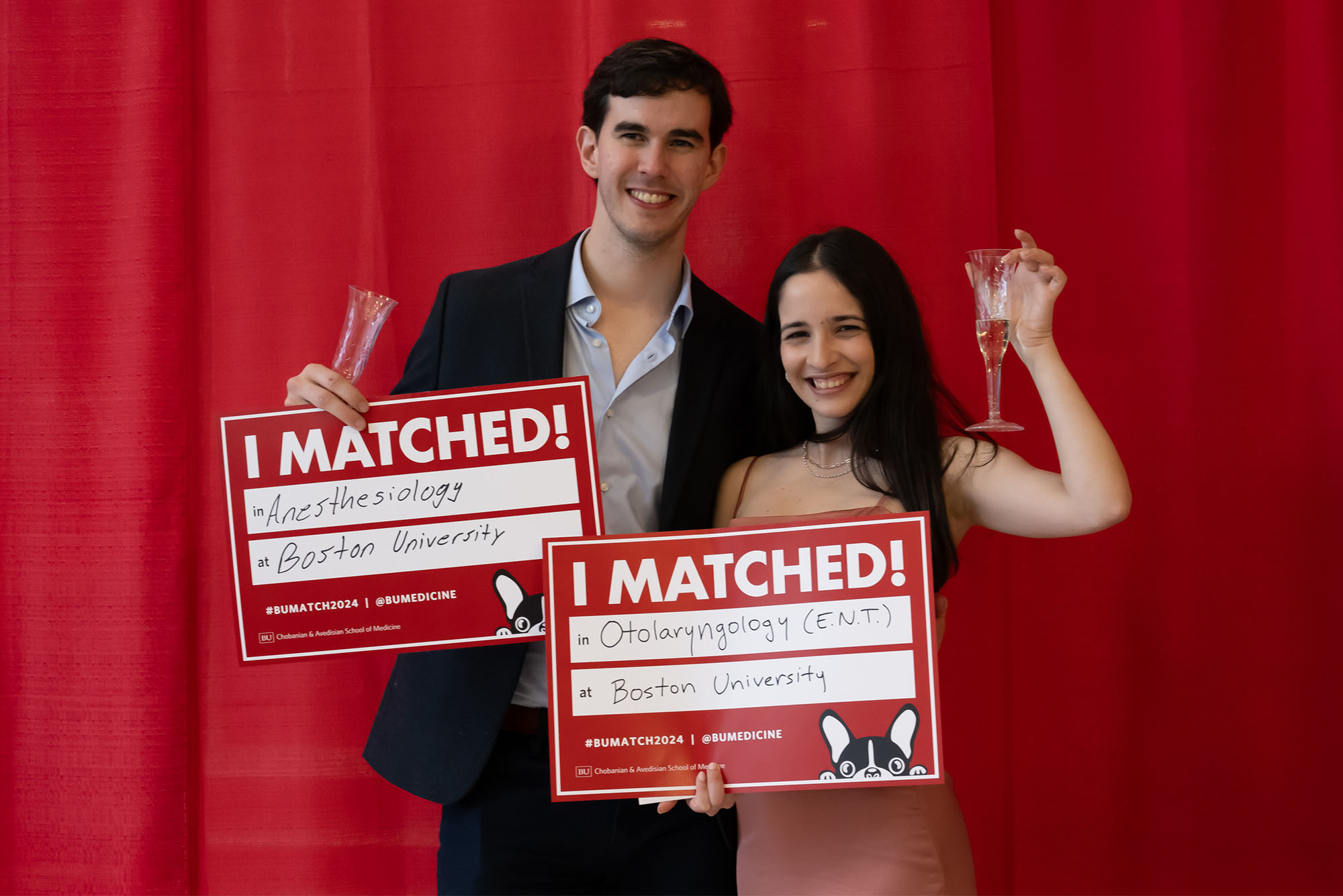 Photo: Two college students in formal attire hold up champagne flutes and signs that read "I Matched! Anesthesiology and Otolaryngology (ENT) @ Boston University" in front of a red background