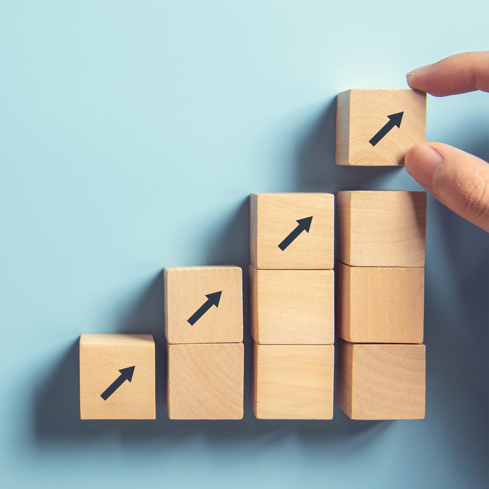 Photo: a hand stacks blocks with arrows pointing upwards on a blue background