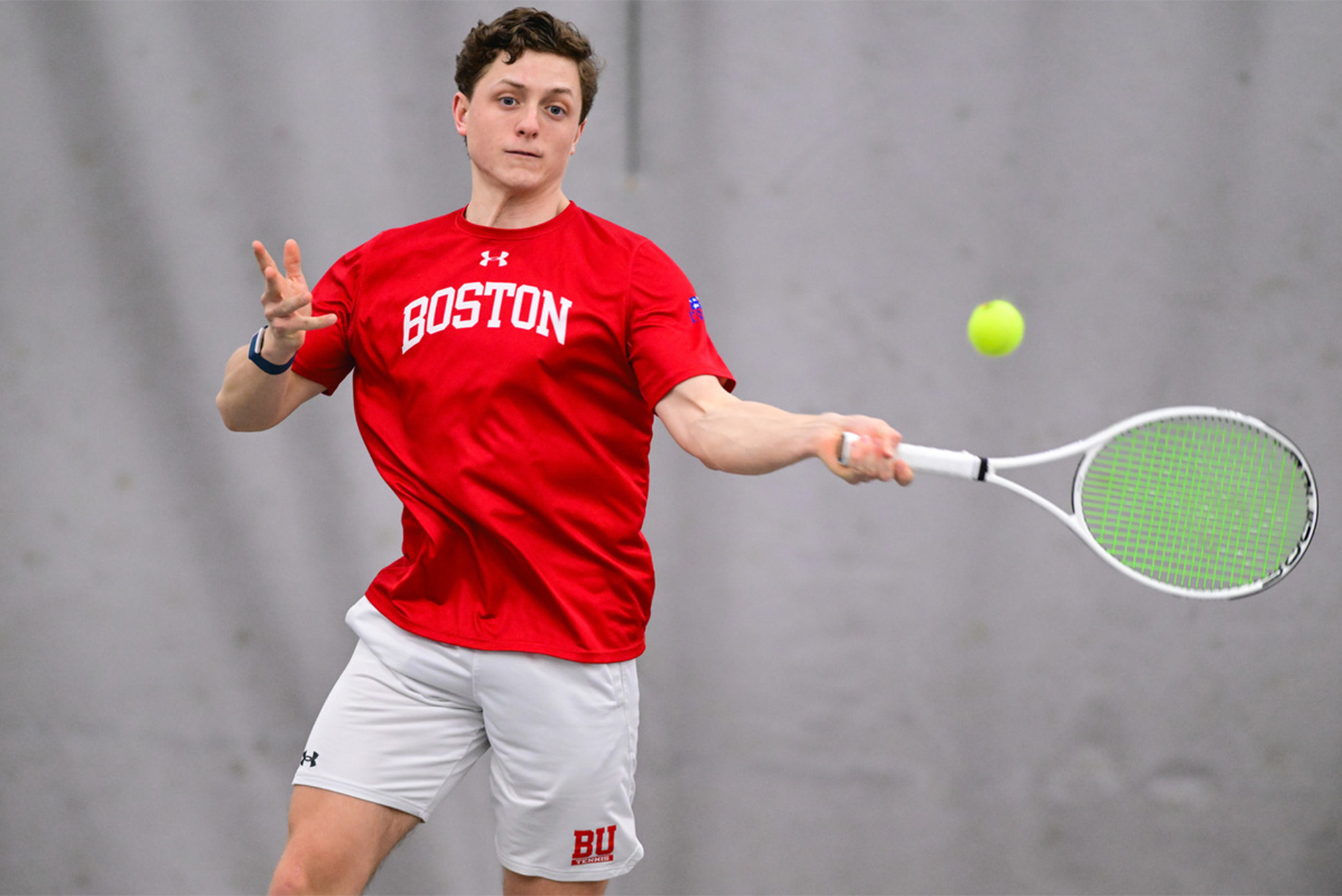 Photo: A college tennis player wearing a red Boston University shirt and white shorts hits a tennis ball at a recent match