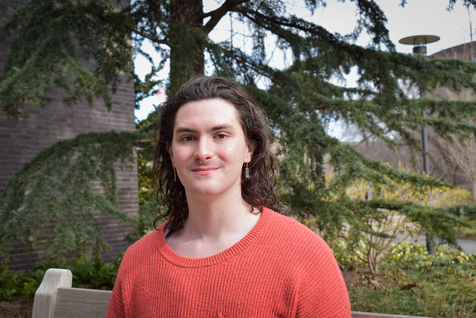 Photo: A person with short dark hair and a nose ring wearing an orange shirt in front of a verdant nature scene with pine trees