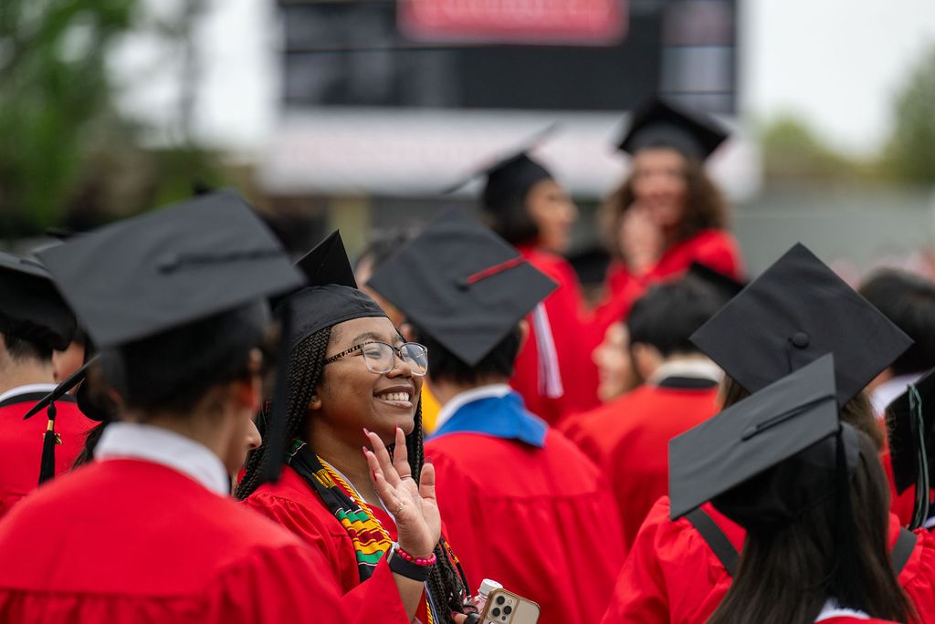 Photo: A picture of many students in black graduation caps and red gowns. One student is smiling and waving in the center of the image