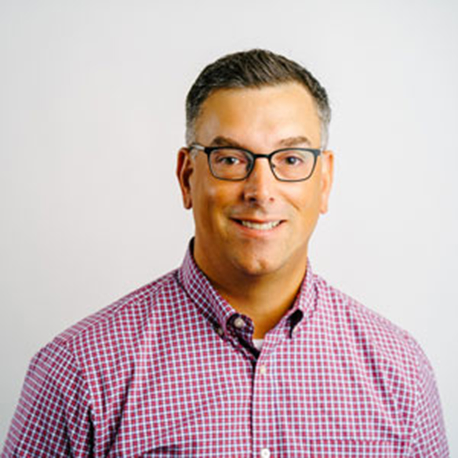 Photo: A headshot of a man with a checkered red shirt. He is wearing glasses and smiling at the camera
