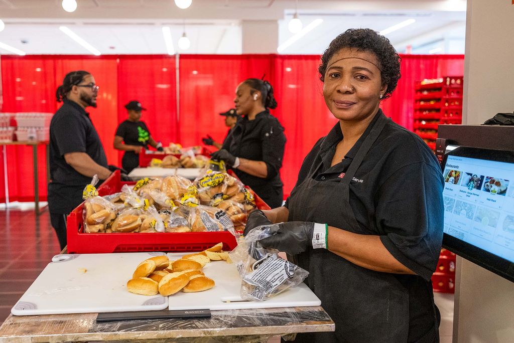 Photo: A picture of a woman smiling softly for the camera. She is wearing a hair net and gloves and seems to be unwrapping packages of rolls