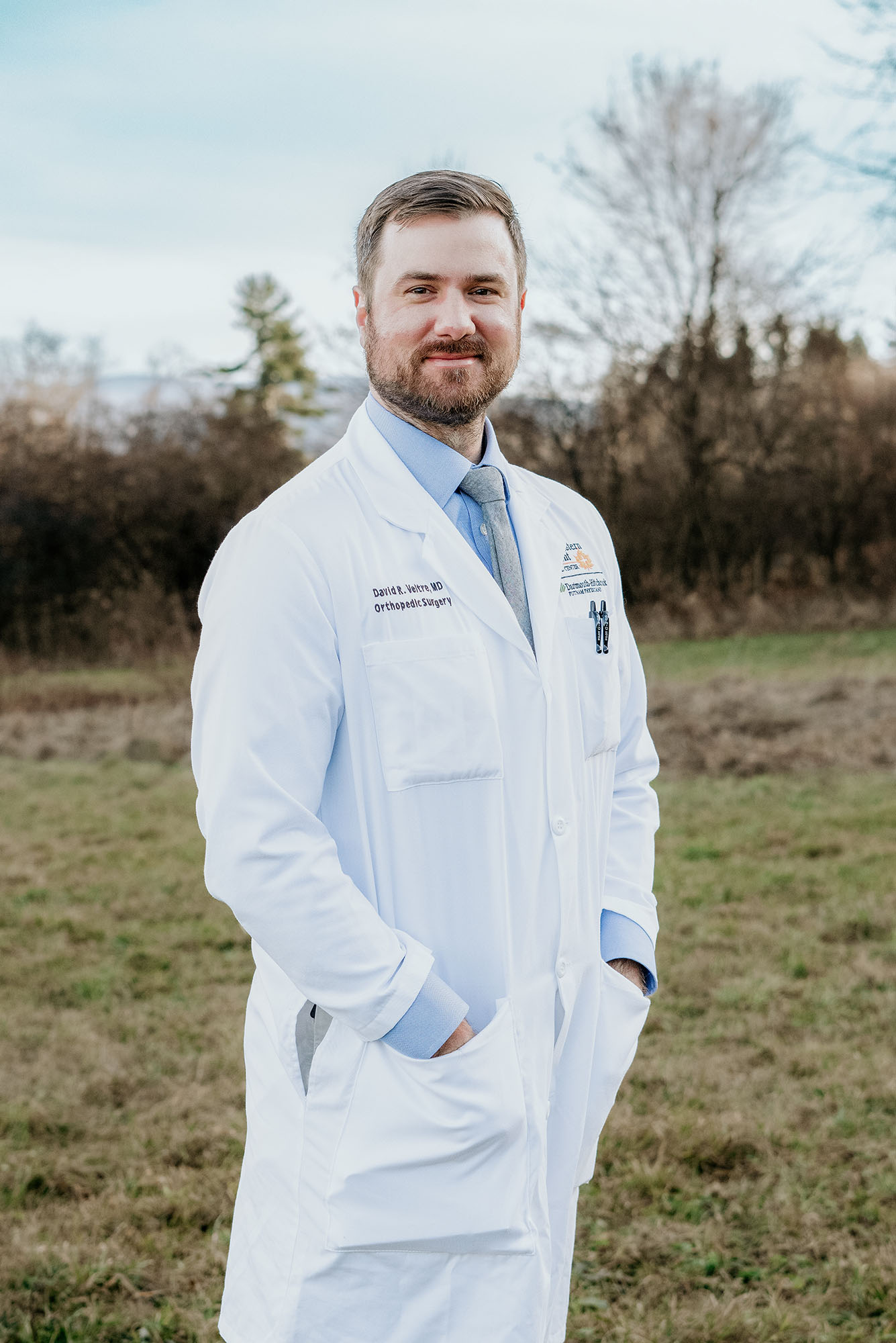 Photo: A white man in a doctor white coat with his hands in his pockets, smiling outside.
