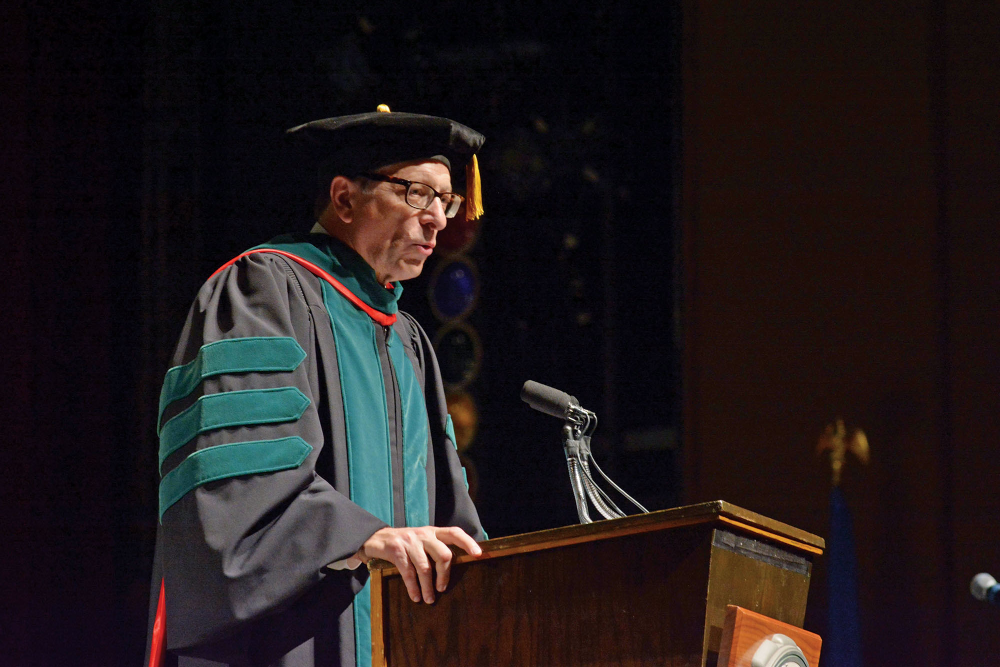 Photo: A old white man with graduation regalia stands at a podium giving a speech.