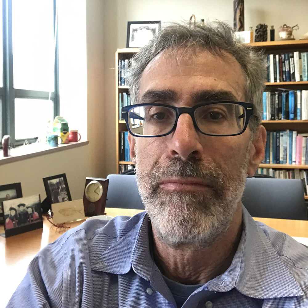 Photo: A man in a button up shirt and black glasses frames takes a selfie. A book shelf stands behind him.