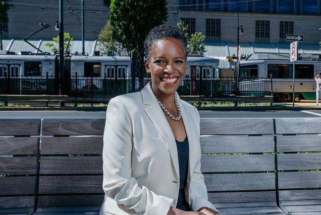 Photo: Boston University's new president Melissa Gilliam, a black woman with short dark hair in a formal outfit, sitting on a wooden bench and a smile in front of an MBTA train stop, in Boston on a sunny day.