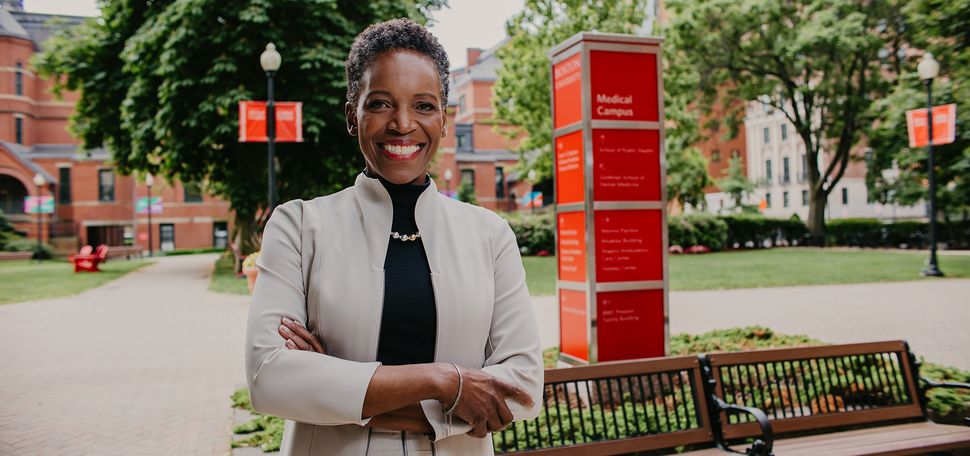 Photo: Boston University's new president Melissa Gilliam, a black woman with short dark hair in a formal outfit stands with arms crossed and a smile in front of a large red Boston University sign, in Boston on a sunny day.