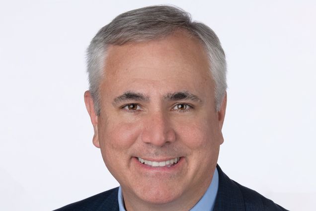 Photo: Lloyd Jones, a white man with short hair and a smile, wearing a navy suit with elegant tie and collared shirt. He is wearing a pin that is a red "8" and a caduceus pin