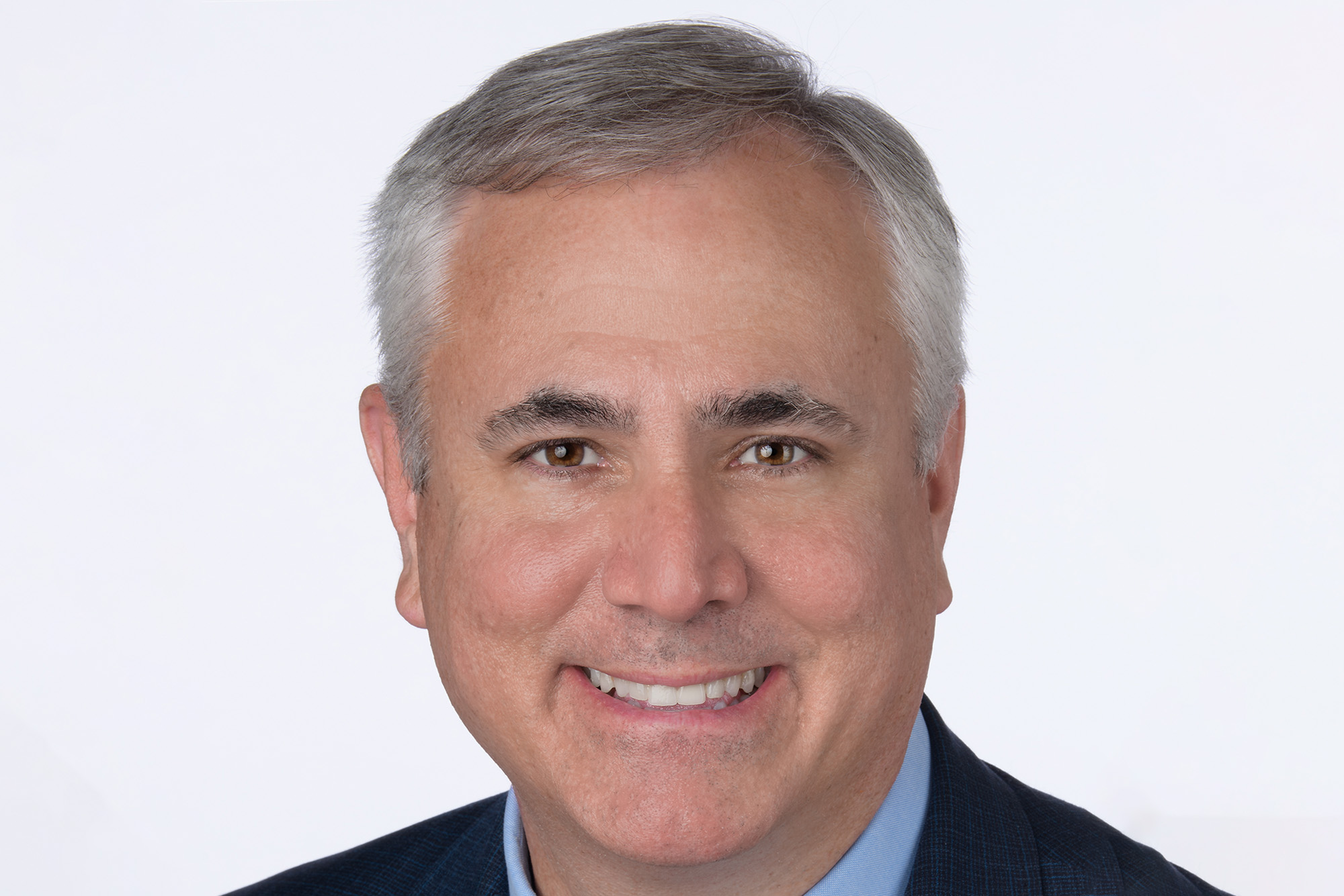 Photo: Lloyd Jones, a white man with short hair and a smile, wearing a navy suit with elegant tie and collared shirt. He is wearing a pin that is a red "8" and a caduceus pin