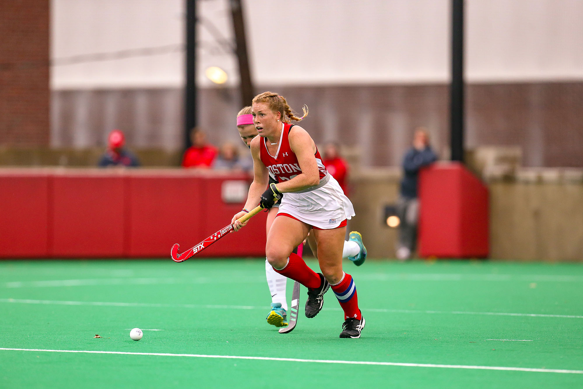 Photo: A white woman running with a field hockey stick on an indoor track.