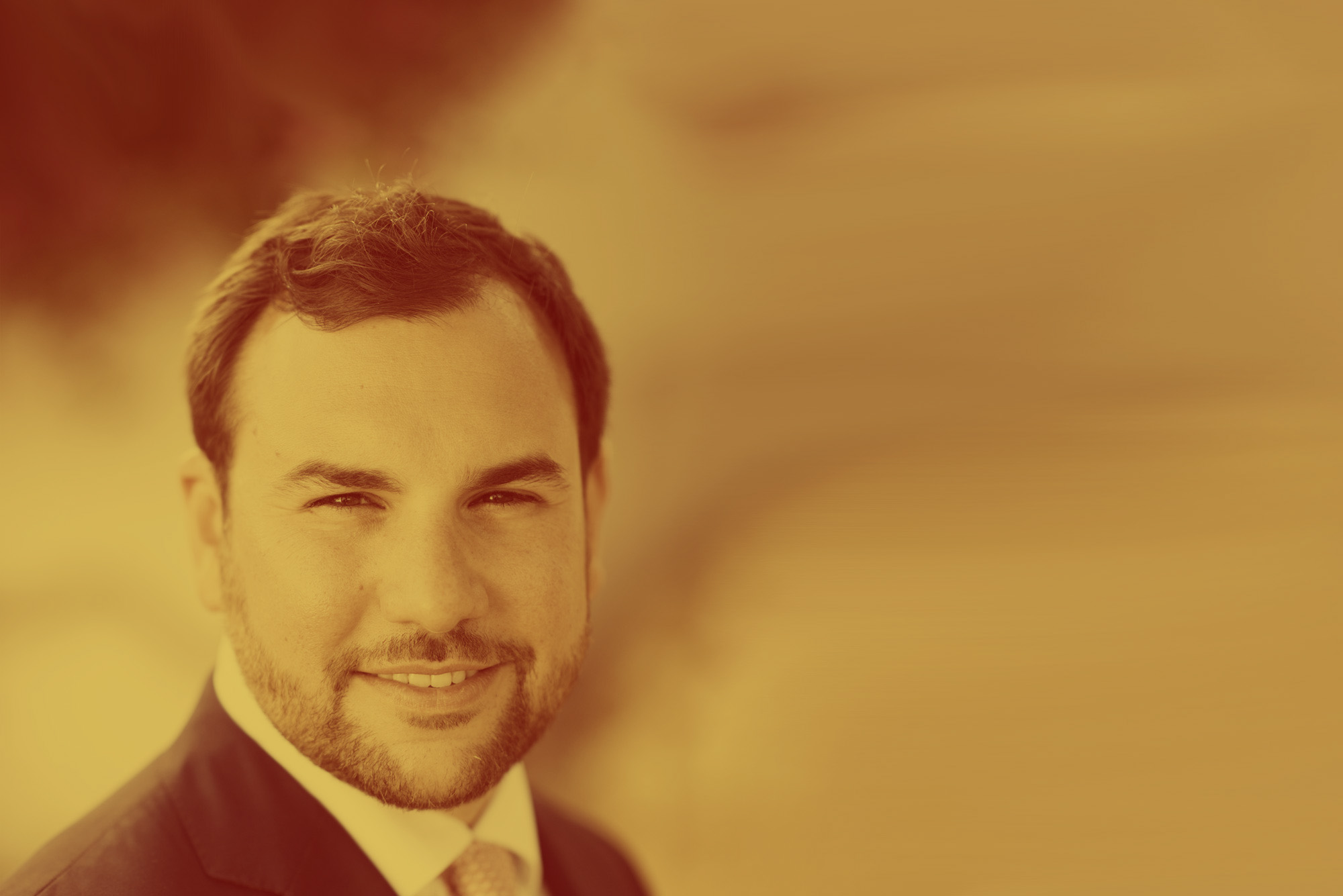 Photo: Formal headshot portrait of a white man with dark hair and beard, wearing a navy suit and white collared shirt. The entire image has an orange colored overlay