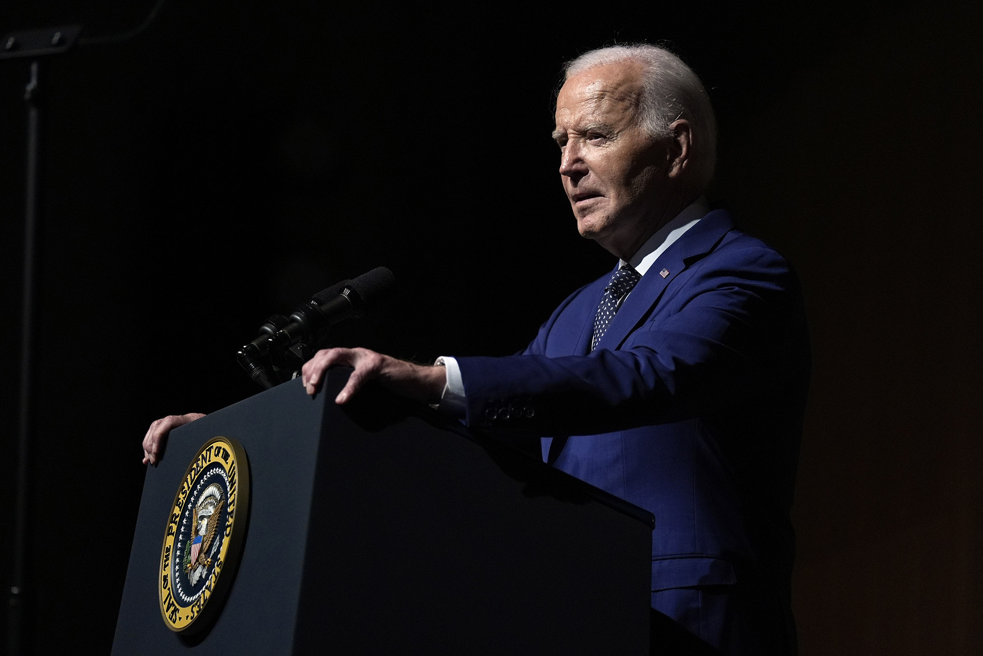 Photo: President Joe Biden speaks at an event. he stands at a podium as the right side of his face is illuminated.