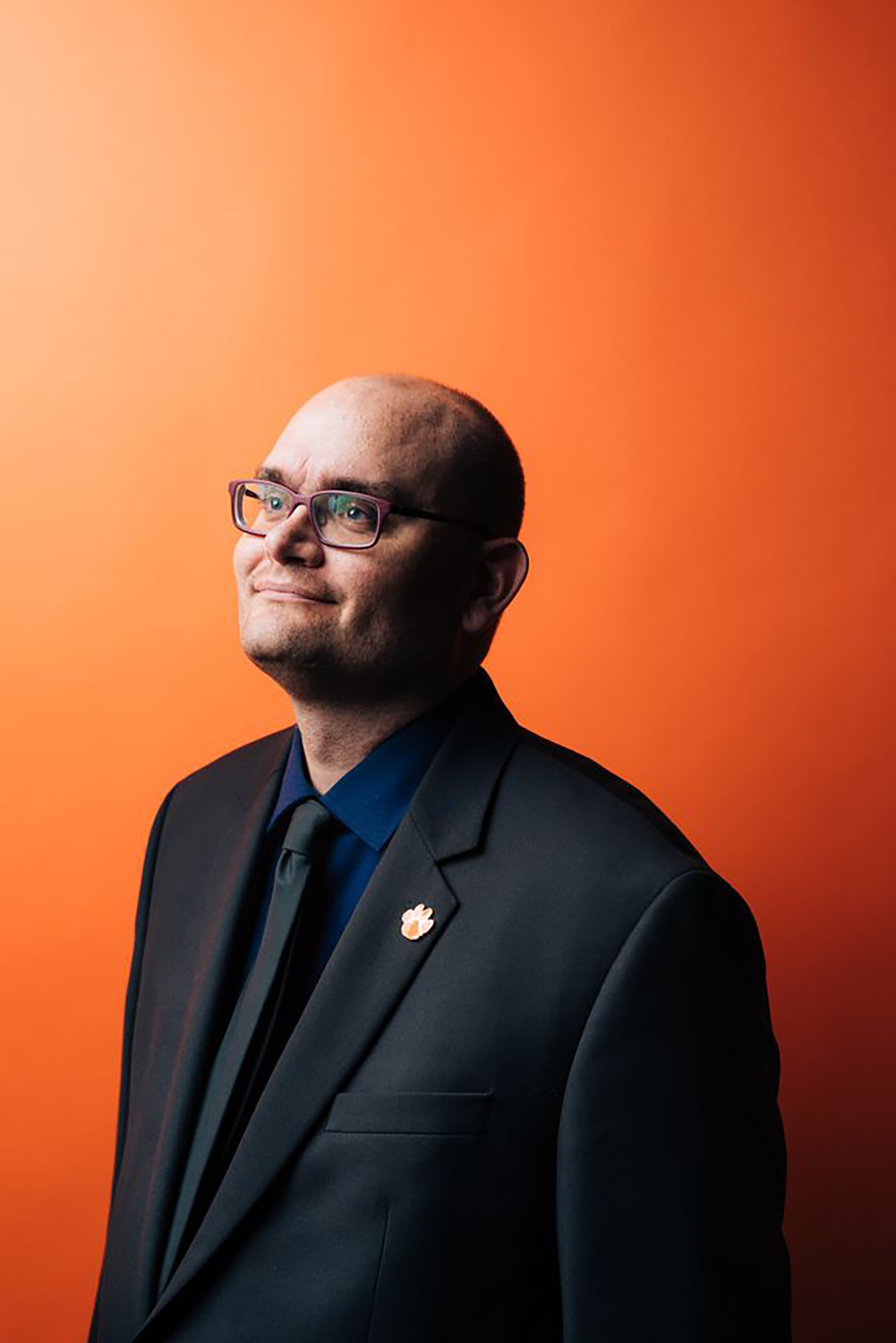Photo: A bald man looks off to the left with maroon glasses while wearing a dark suit. He stands in front of a orange background.