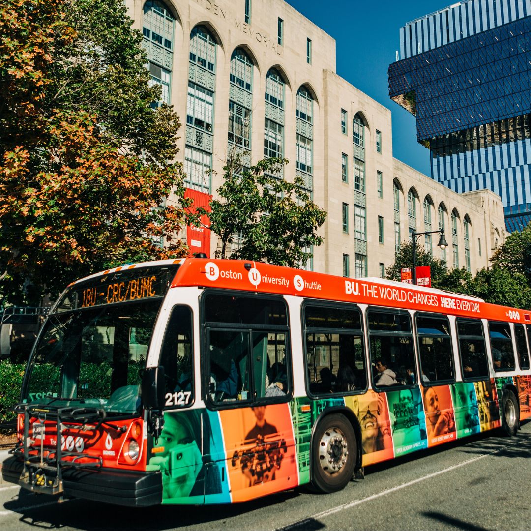 Photo: The Boston University BUS (BU Shuttle), a red and white University branded shuttle bus, driving down Commonwealth Avenue on a sunny day. The background features the University's College of Arts and Sciences building, and trees with leaves that are starting to change color.