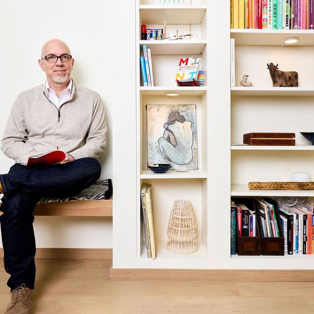 Photo: A bald man with a goatee sits next to a bookcase filled with art and books