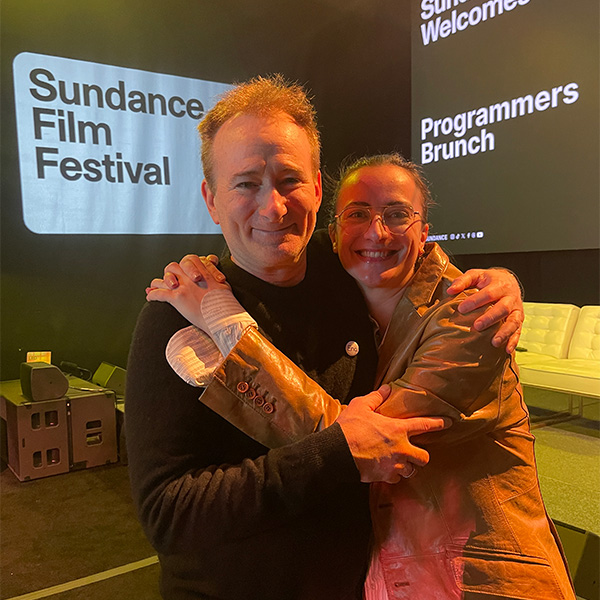 Photo: A man and woman hug each other in front of a white banner with black text reading "Sundance Film Festival"