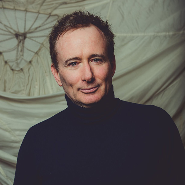 Photo: A white man in a turtleneck poses for an artisy headshot in front of a grey background