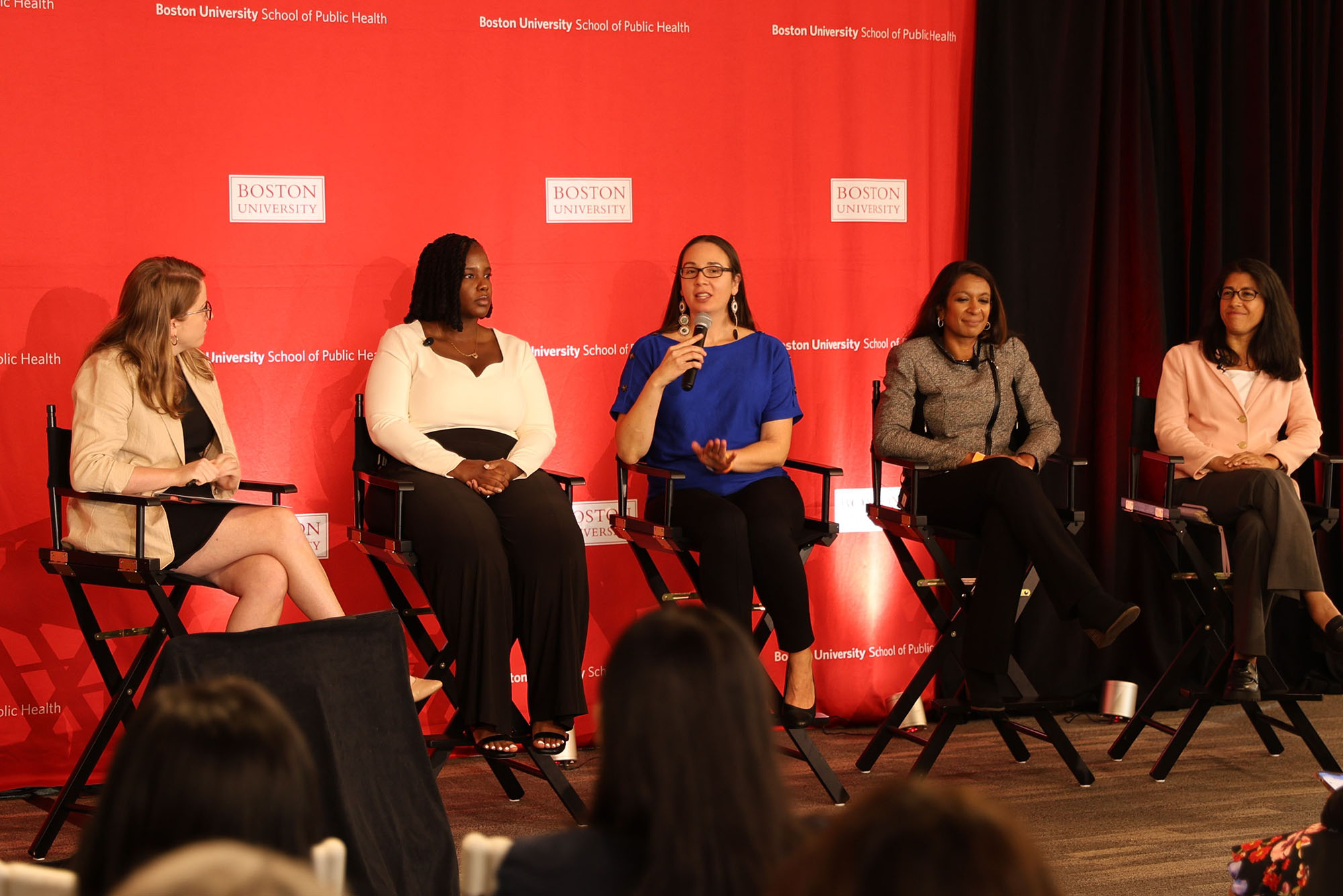 Photo:A picture of people sitting on chairs on a stage during an event. Behind them is a red wall that has the "Boston University" logo placed on it many times. There are people in chairs in the audience looking up at them