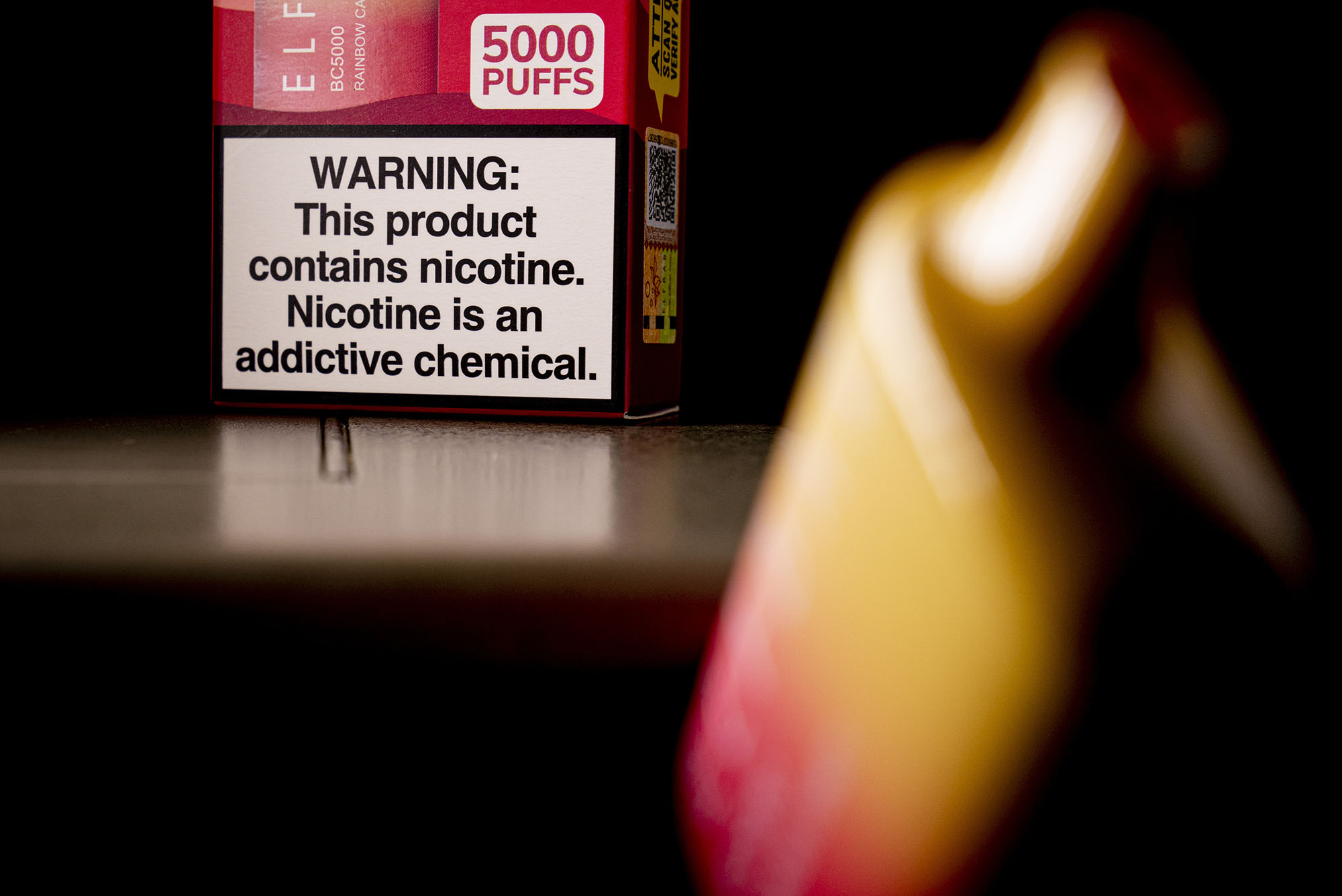 Photo: An Elf Bar disposable vaping pod device is displayed in the foreground, blurred. Behind the vape stands the packaging with a big warning label at the bottom.