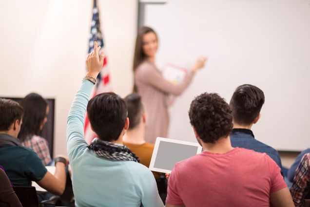 Photo: A student raising his hand during a class. His female teacher is turning to look back at him and stands at the front of a whiteboard next to an American flag