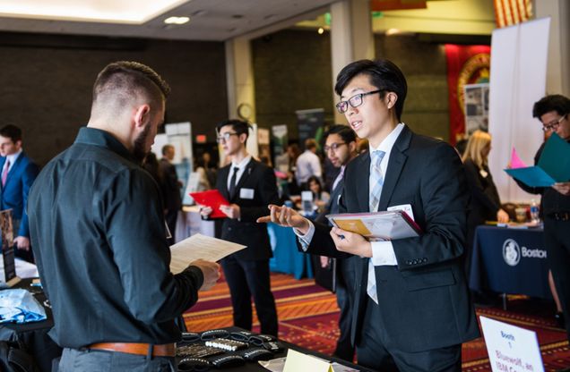 Photo: A busy job fair scene where several people dressed in business attire are conversing. In the foreground, a young man in a suit and tie, holding a folder and documents, is actively speaking with another man wearing a dark shirt who is holding a paper. Other individuals in the background, also in suits, are interacting and exploring the event booths, which have banners and materials set up for recruitment. The event appears to be professional and career-focused, taking place in a large, well-lit room.