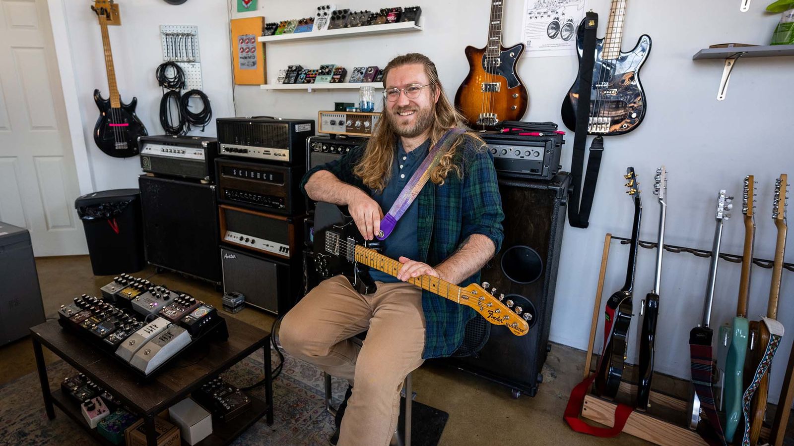 Photo: A picture of a man with long hair wearing a green and blue flannel sittingo n a chair and holding a guitar. He is surrounded by other guitars and guitar pedals