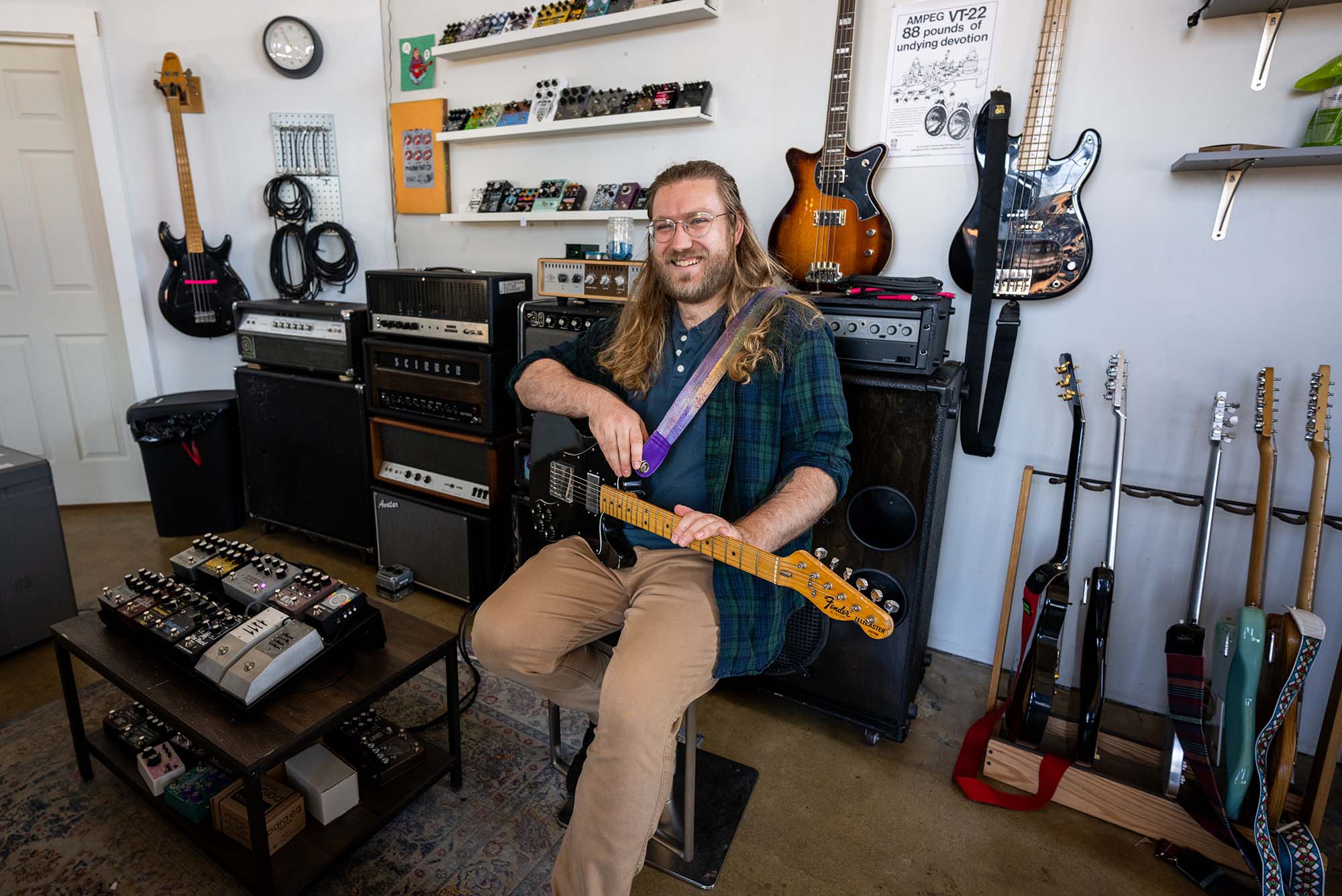 Photo: A picture of a man with long hair wearing a green and blue flannel sittingo n a chair and holding a guitar. He is surrounded by other guitars and guitar pedals