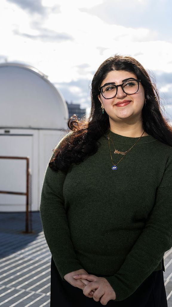Photo: A female student standing on a rooftop with a closed smile, looking off in the distance