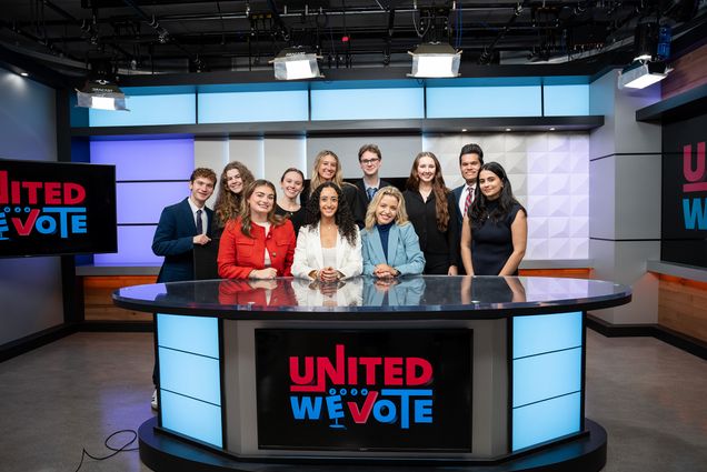 Photo: A news desk with a group of students posing behind it with the words "United We Vote" on the front of it