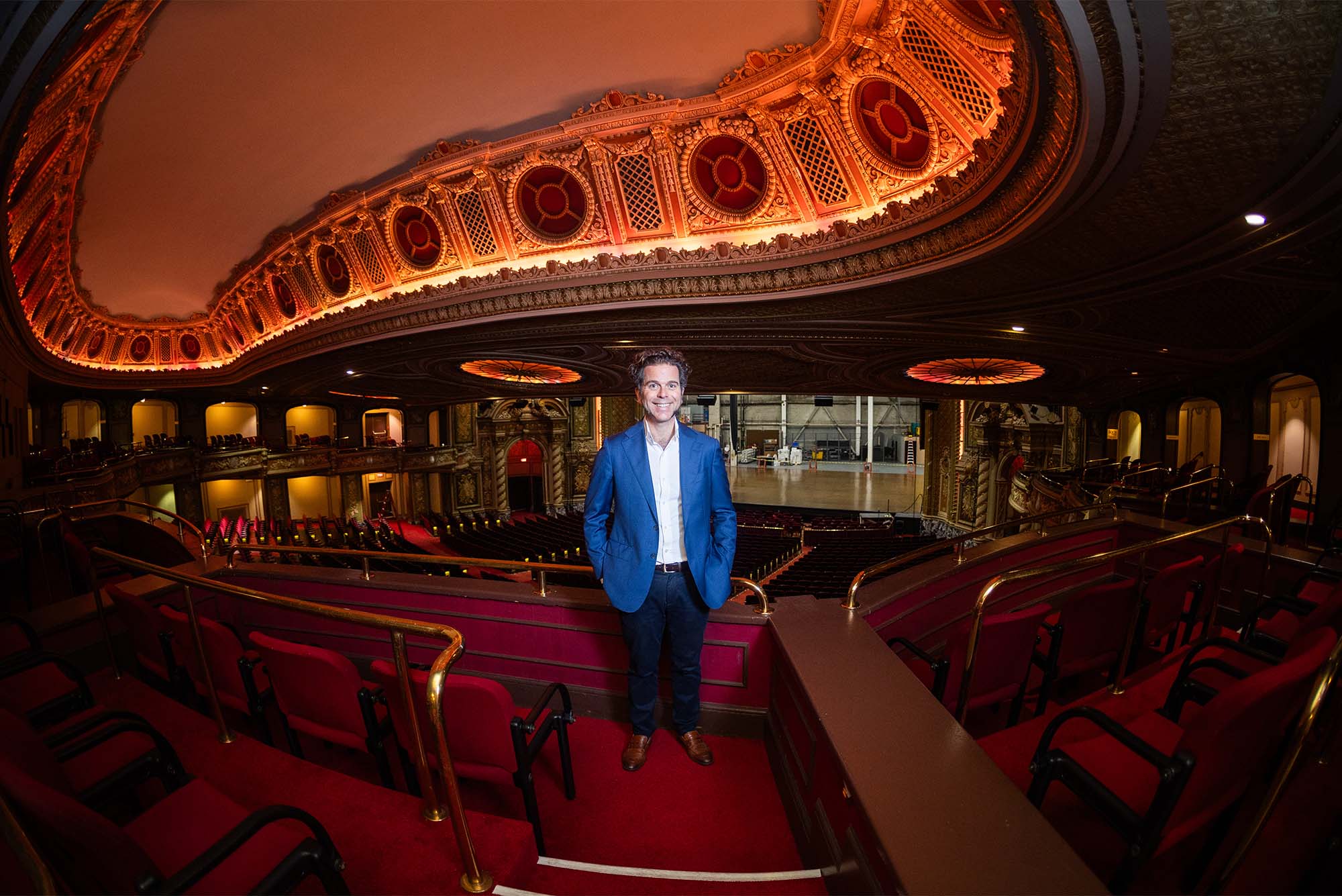 Photo: A man in a blue suit stands inside an elegant theatre lit with warm lighting
