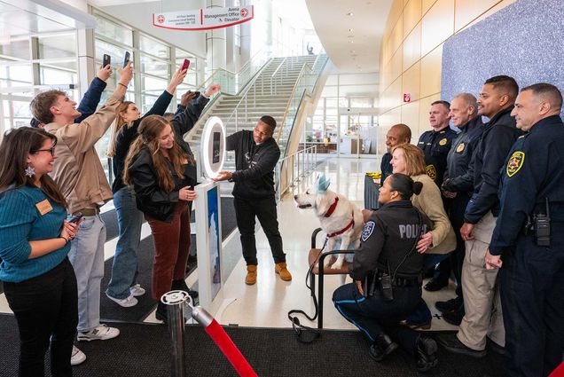 Photo: Bean, a golden retriever, in front of a photo booth with members of BUPD surrounding her. Onlookers are taking paparazzi-style photos of the comfort dog as well