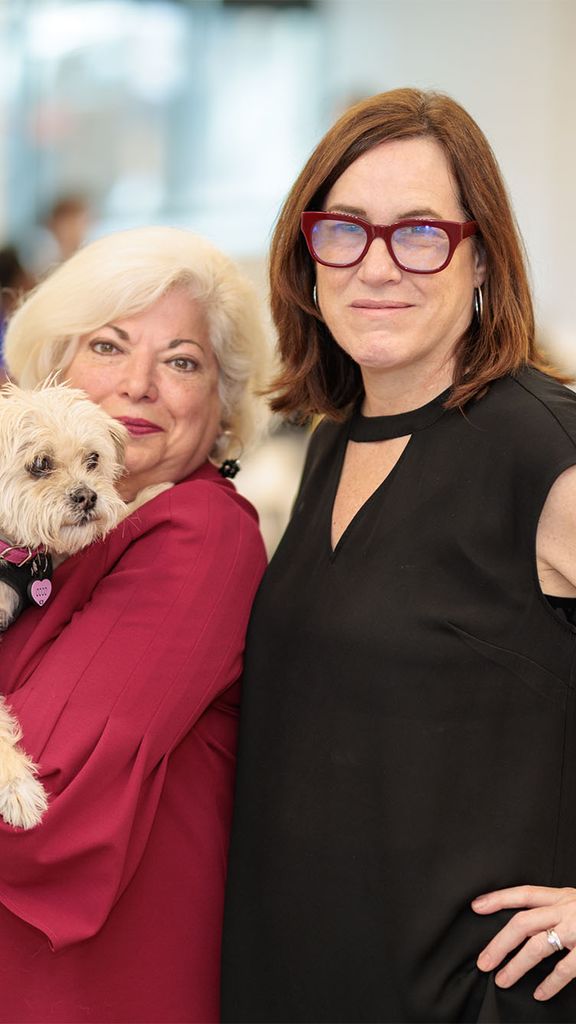 Photo: Two women standing side by side, one wearing red and holding a small white dog, the other wearing black with black glasses