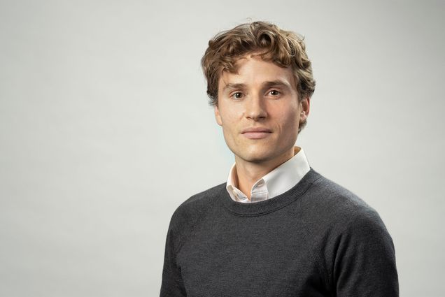 Photo: Man in a collared white shirt and dark sweater poses for a formal headshot in front of a light background