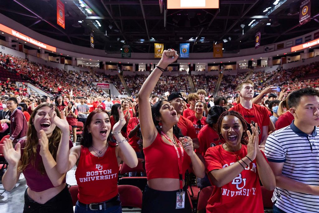 A crowd of BU tailgaters dressed in red, BU merchandise cheer in unison. The crowd is in a domed, sports arena with bright lights.