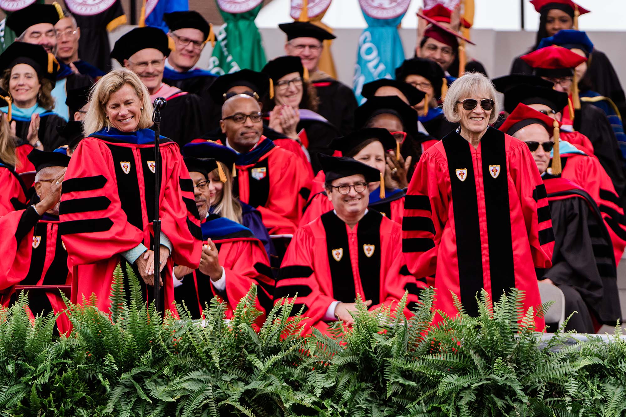 Photo: People standing on a stage lined with greenery in Boston University red academic robes with black trim and caps