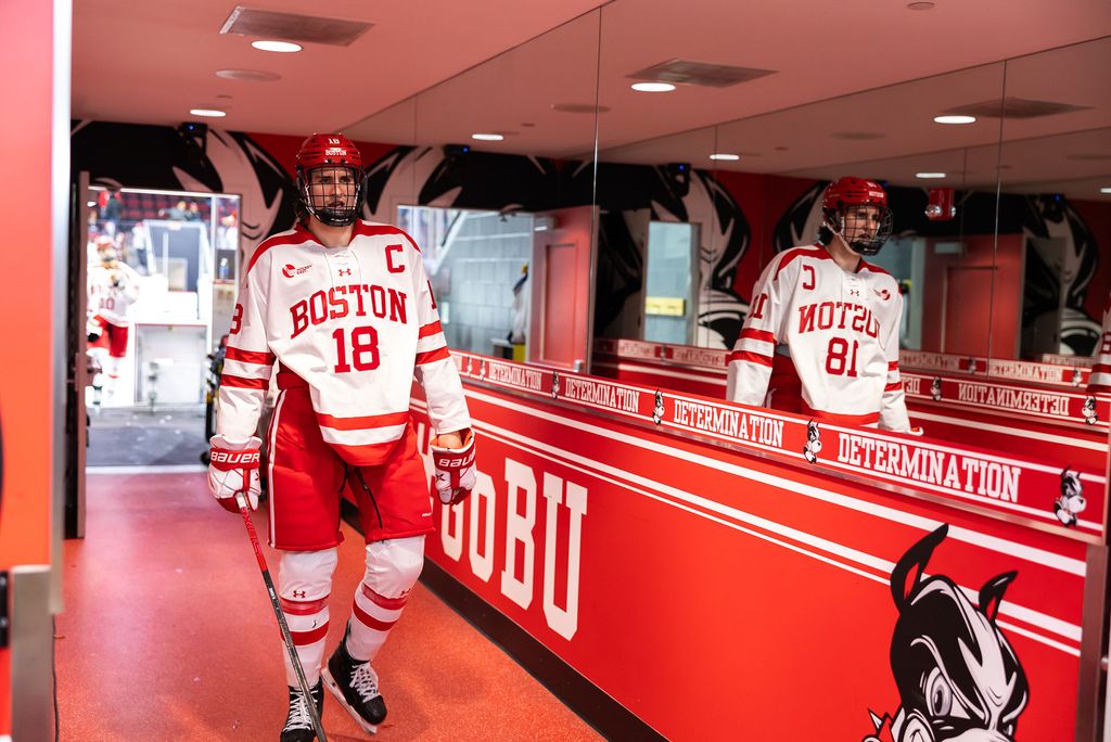 Photo: A hockey player wearing a Boston University uniform and walking down a red hallway with images of BU's terrier mascot along it