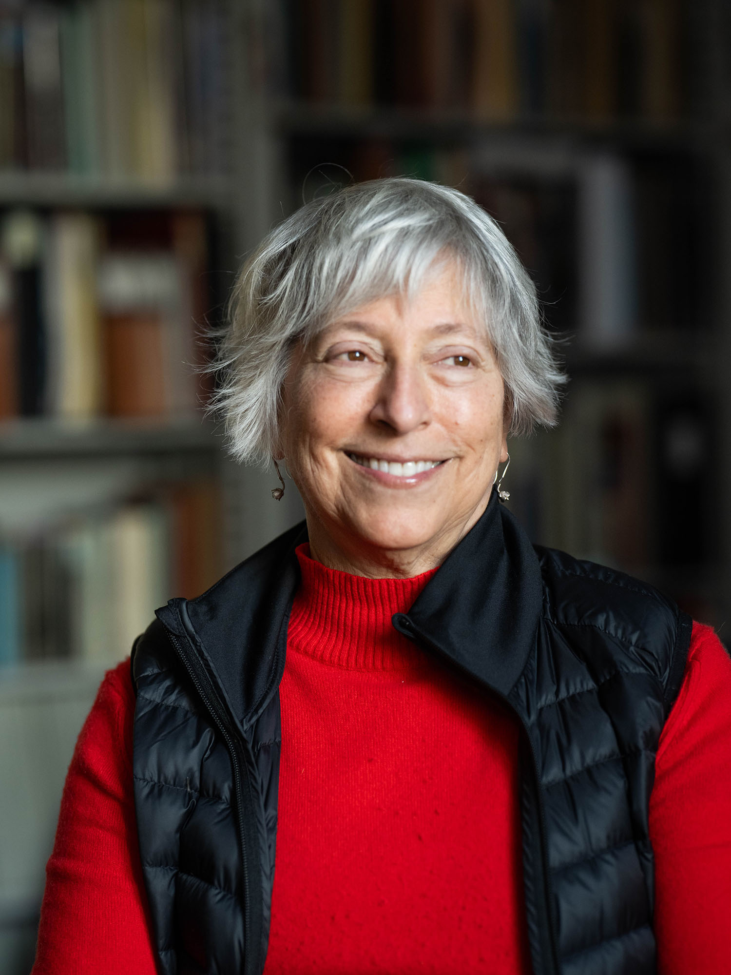 Photo: A woman with gray, short hair smiling and looking off to the side. She is wearing a red turtleneck sweater with a black vest