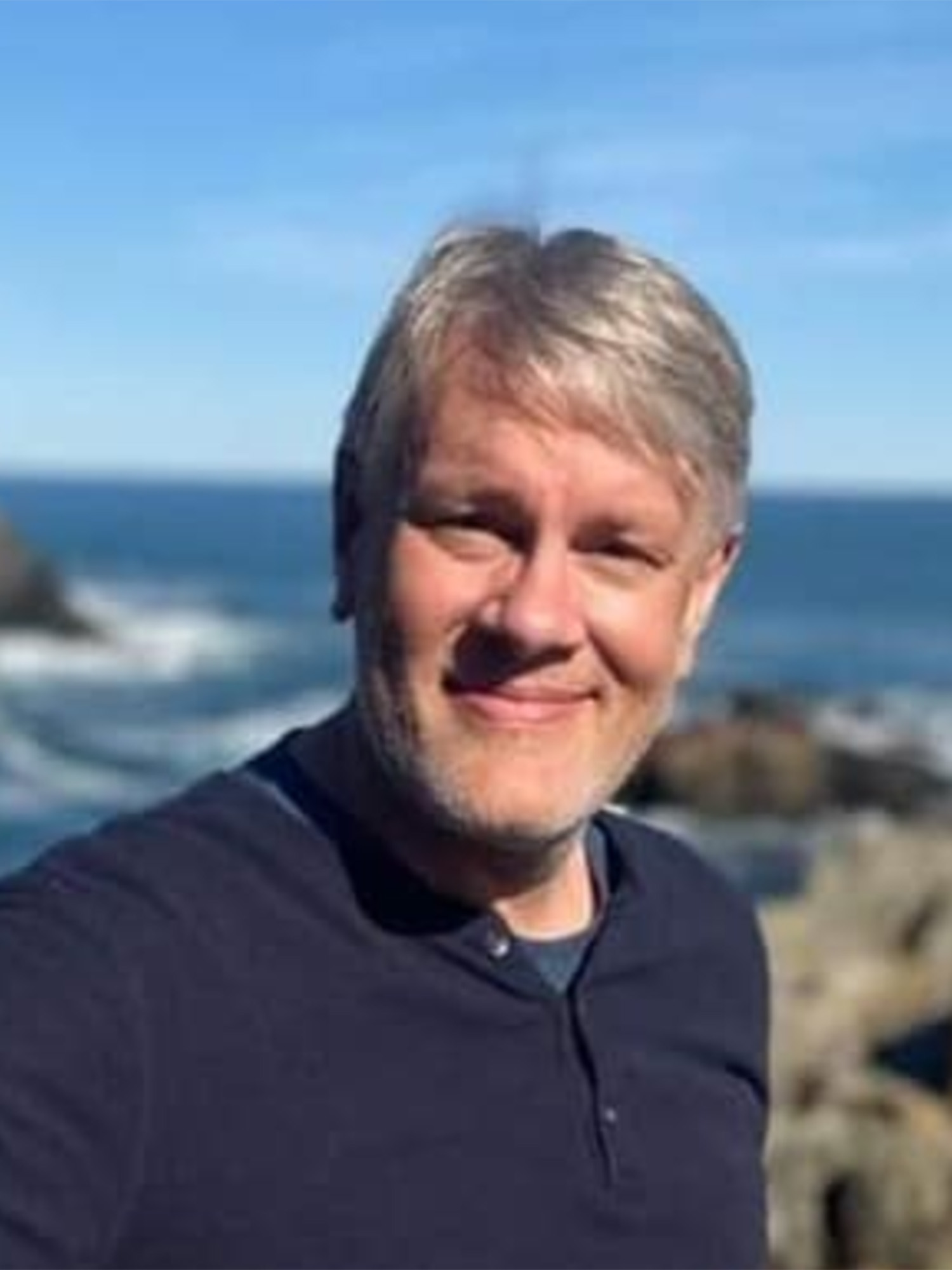 Photo: A headshot of a man with gray hair in a blue shirt on a rocky beach