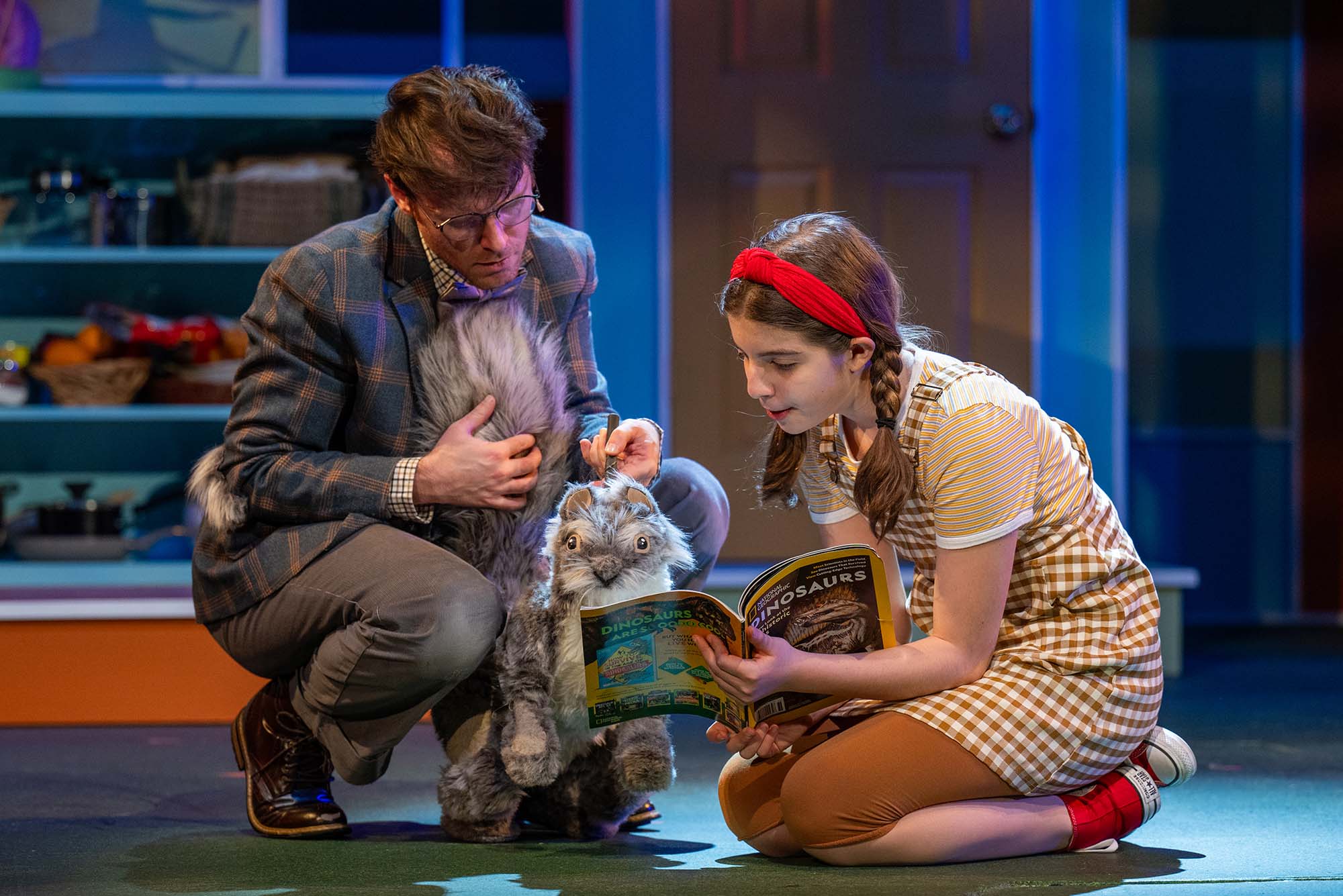 Photo: A man on the left controlling a squirrel puppet as a young girl on the right shows it an open magazine