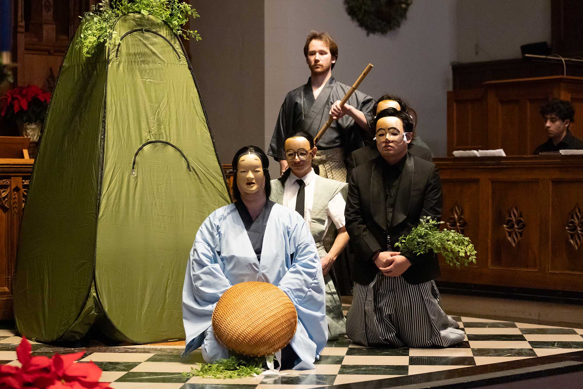 Photo: Individuals on a stage presenting a traditional Japanese Noh. In the middle of the stage, a individual rest on their knees. Behind them, two more people kneel. And behind them, a man standing with a staff.