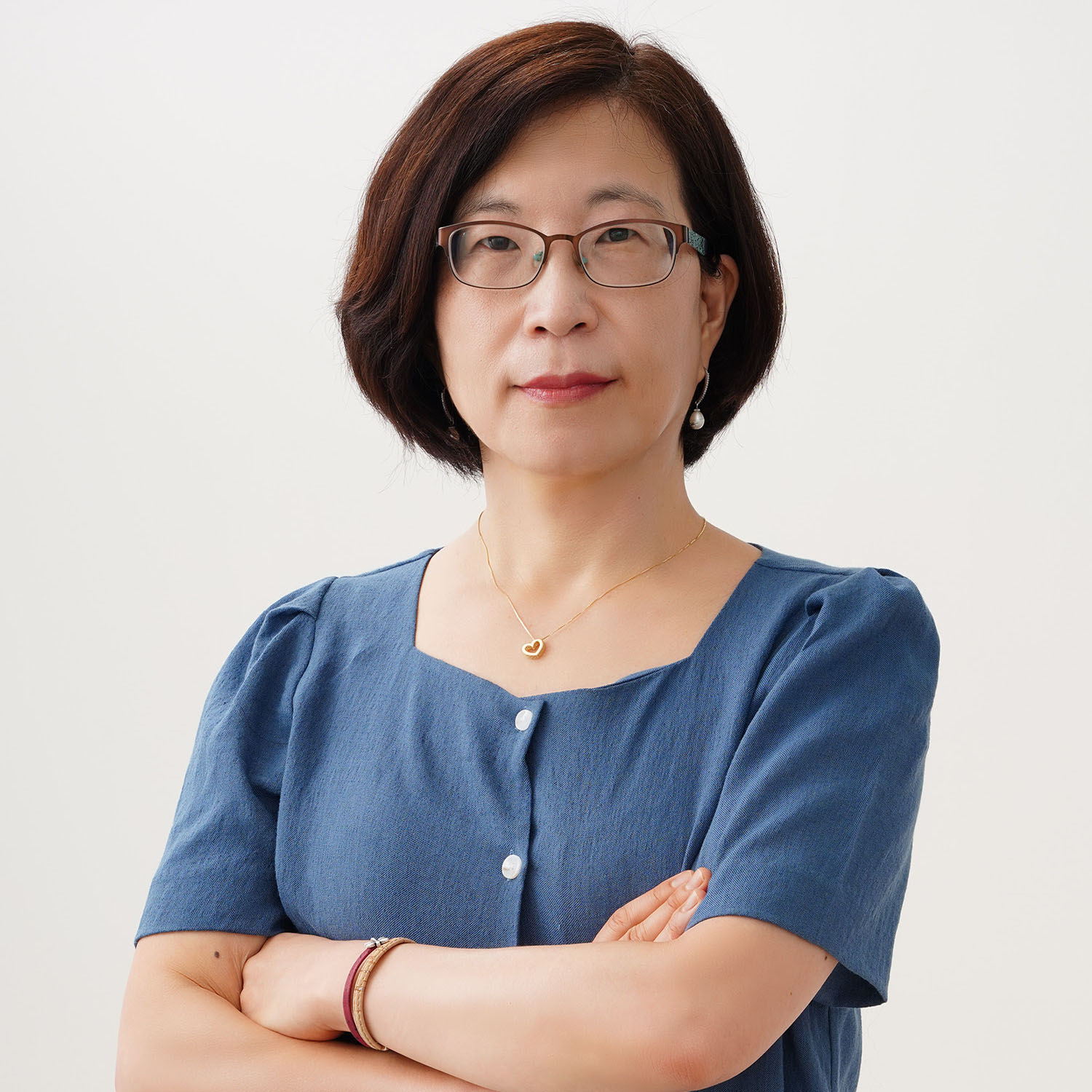 Photo: A headshot of a Korean woman with short hair and glasses. She is wearing a blue, short sleeved button up shirt and a gold heart necklace