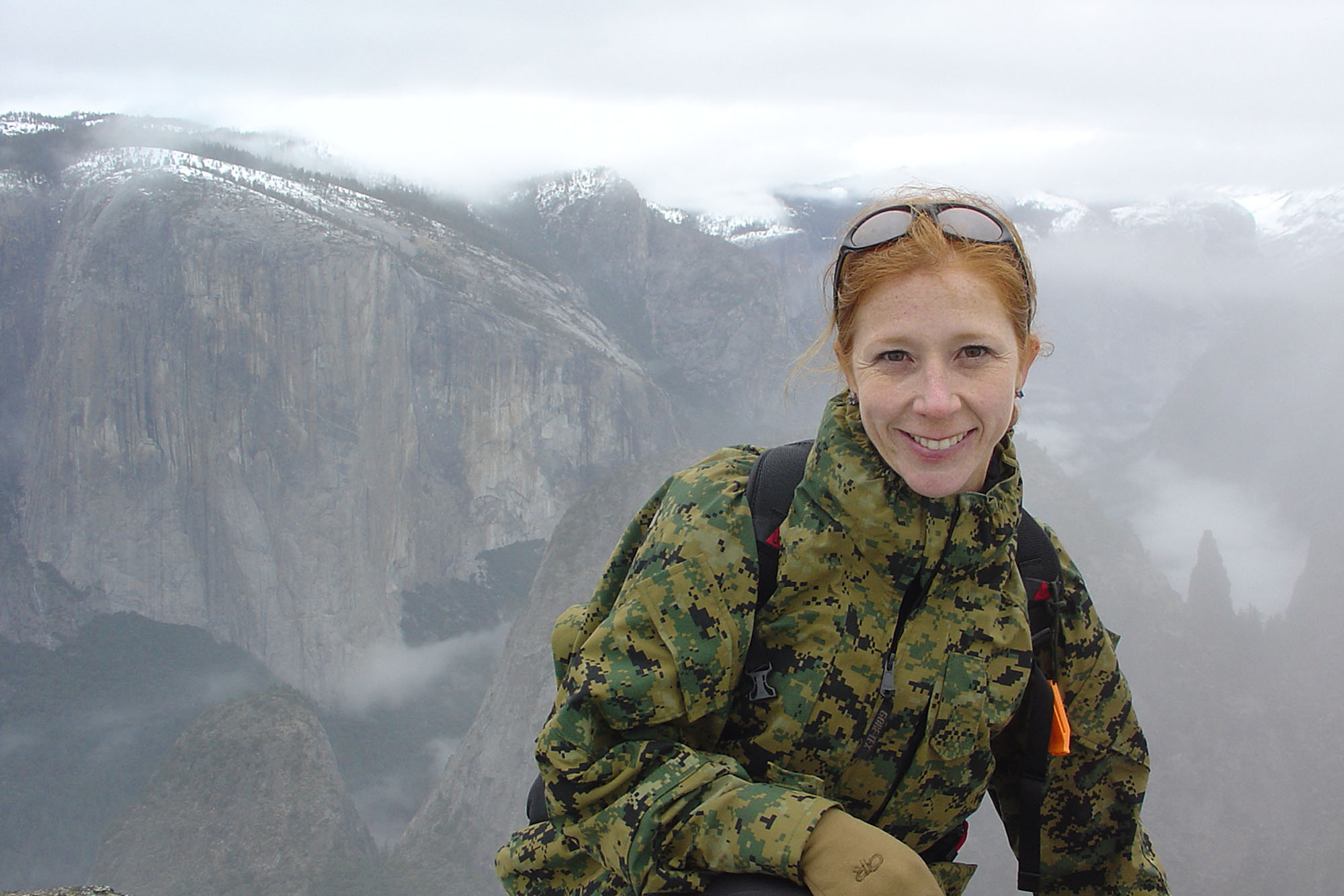 Photo: A woman with red hair smiling and posing in front of a snowy mountain landscape while wearing a camo jacket