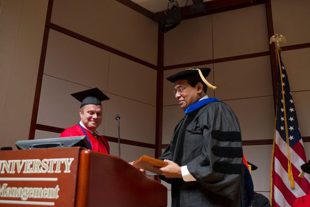 Photo: Two men on stage. The one on the left is wearing a red gown and cap and the man on the right is wearing a black gown and cap